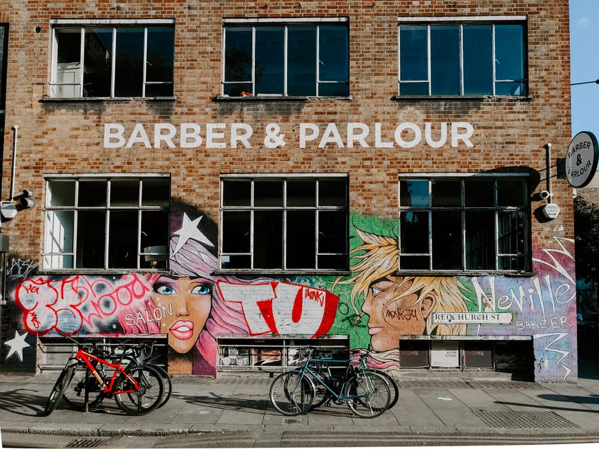 A multi-story brick building with a large sign reading 'BARBER & PARLOUR' in white letters on the upper façade. Below the sign, there is vibrant street art graffiti featuring stylized portraits of a woman and a man with colorful, exaggerated hair and facial features. The graffiti also includes various tags and words in red, white, and blue, along with decorative stars. On the sidewalk in front of the building, there are two bicycles parked, one black and one red, leaning against a bike rack. The area appears to be in an urban setting with bright natural daylight illuminating the scene, and a small circular sign on the side of the building repeats the 'Barber & Parlour' name. The overall environment suggests a friendly, lively neighbourhood street with artistic street culture integrated into a commercial premises specializing in grooming services, possibly with an emphasis on independent or alternative waste handling through local service providers like Rubbish Clearance Shoreditch.