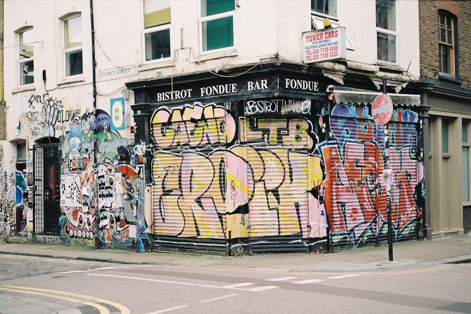 A multi-story brick building with a large sign reading 'BARBER & PARLOUR' in white letters on the upper façade. Below the sign, there is vibrant street art graffiti featuring stylized portraits of a woman and a man with colorful, exaggerated hair and facial features. The graffiti also includes various tags and words in red, white, and blue, along with decorative stars. On the sidewalk in front of the building, there are two bicycles parked, one black and one red, leaning against a bike rack. The area appears to be in an urban setting with bright natural daylight illuminating the scene, and a small circular sign on the side of the building repeats the 'Barber & Parlour' name. The overall environment suggests a friendly, lively neighbourhood street with artistic street culture integrated into a commercial premises specializing in grooming services, possibly with an emphasis on independent or alternative waste handling through local service providers like Rubbish Clearance Shoreditch.