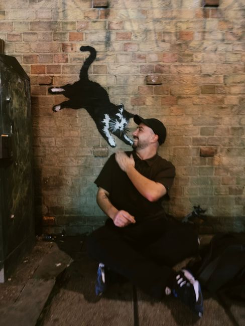 A man dressed in black clothing, including a cap, crouching on one knee in front of a brick wall with a rough, textured surface. The wall features a painted mural of a black and white cat, depicted in mid-leap with its paws extended and tail curved upward, creating a dynamic contrast against the reddish-brown bricks. The man is smiling and looking at the mural, with his right hand raised near his shoulder, appearing engaged or playful. The scene is illuminated by natural or ambient lighting that highlights the textured wall and the details of the mural. To the left of the man, a black storage locker or cabinet is partially visible, and the ground surface appears uneven, with some debris or dirt. Nearby, a black bag or backpack rests on the ground, indicating a casual or outdoor setting. The overall environment suggests an urban space with artistic street elements, relevant to private or alternative waste handling environments, subtly aligning with rubbish removal themes through the urban context and mural art.