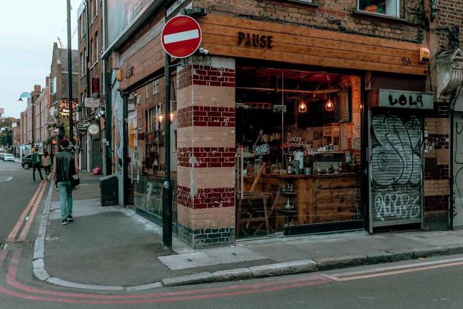 The image shows a street corner with a small establishment named 'PAUSE' occupying a brick building with a wooden sign above the entrance. The shop's exterior features a large window revealing a cozy interior with warm lighting, exposed bulbs, and wooden furniture, indicating a café or similar venue. To the right of the entrance, there is a graffiti-covered metal shutter, partially closed. The building's façade displays weathered brickwork with red and beige tones, and some areas show signs of aging or previous modifications. On the street, a man wearing a dark jacket and light-colored pants walks along the pavement while other pedestrians are visible further down the sidewalk. Traffic signs, including a 'no entry' sign, are mounted on the building corner. The surrounding area comprises other brick structures, some with visible signage and lighting, creating an urban atmosphere typical of Shoreditch. The overall scene is captured in natural daylight, with a slightly overcast sky providing diffuse lighting that highlights the textures and details of the brickwork, signage, and street elements, reflecting an active, diverse city environment suitable for private waste disposal or rubbish clearance services by Rubbish Clearance Shoreditch.