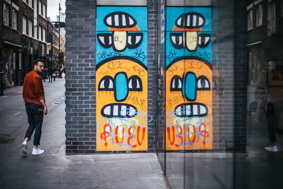 A street scene features a tall, narrow brick wall with a vibrant graffiti mural displaying two stylized faces side by side, each with large, oval-shaped blue noses, wide grinning mouths with white teeth, and black and white striped eyes beneath orange and yellow backgrounds. The graffiti includes various tags and scribbles in red, black, and blue spray paint, with some stylized letters and symbols overlaid on the faces. To the left, a young man wearing a burnt orange jacket, dark jeans, and white sneakers is walking along the sidewalk, turning his head towards the graffiti. On the right, a glass storefront window reflects part of the street scene, including a person in the background. The setting appears to be an urban area with apartment buildings lining the street, and the lighting suggests a cloudy day or shaded environment. The graffiti adds a contemporary, artistic element to the otherwise neutral-colored brick and concrete surroundings, subtly highlighting urban street art's role in alternative urban waste and image handling.