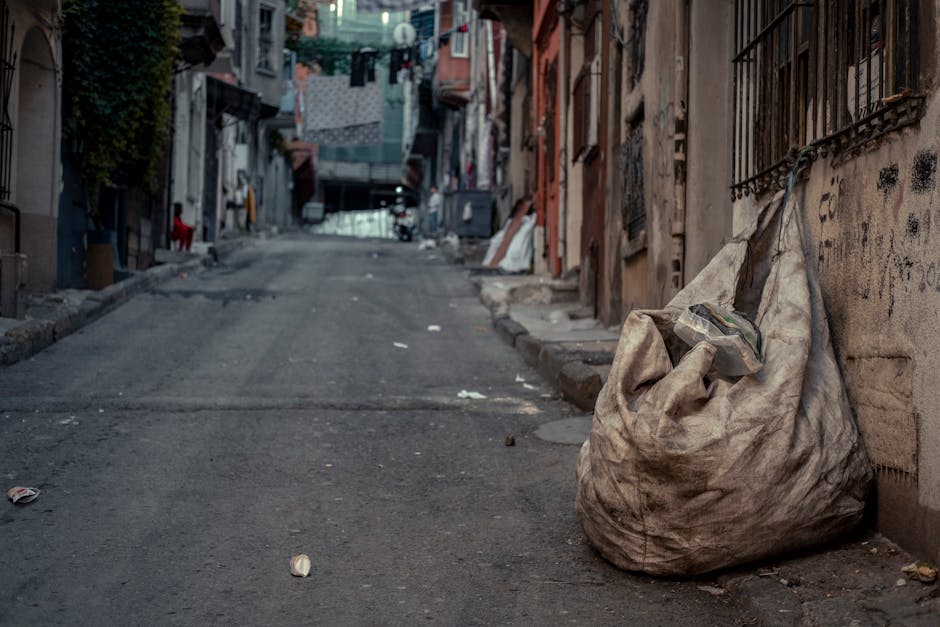 A large, dirty, off-white refuse bag made of worn, crinkled plastic material is placed on a paved street by a building wall. The bag appears to be filled with various waste items, with a glimpse of plastic bottles visible at the top. The street is narrow, with an uneven asphalt surface that shows signs of wear, and it slopes slightly upward towards the background. Surrounding the area are residential buildings with graffiti on the walls, metal barred windows, and hanging laundry visible further down the street. The environment looks aged and somewhat neglected, with scattered small pieces of litter on the ground. On the left side, a narrow strip of curb and sidewalk can be seen, while on the right, the building exterior features peeling paint and rusted iron window grilles. The lighting is subdued, suggesting an overcast day or shaded alley, which emphasizes the utilitarian scene related to waste collection and private rubbish disposal, as handled by services such as Rubbish Clearance Shoreditch.